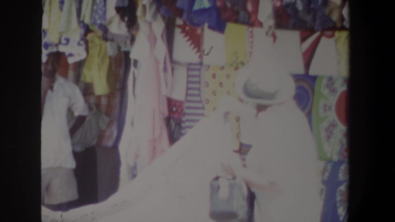 A seller stands behind hanging clothes while a customer in a light hat faces the stall.