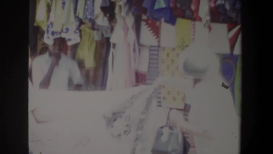 A market seller remains visible behind colorful textiles as the foreground customer and clothing are washed by bright 8mm exposure.