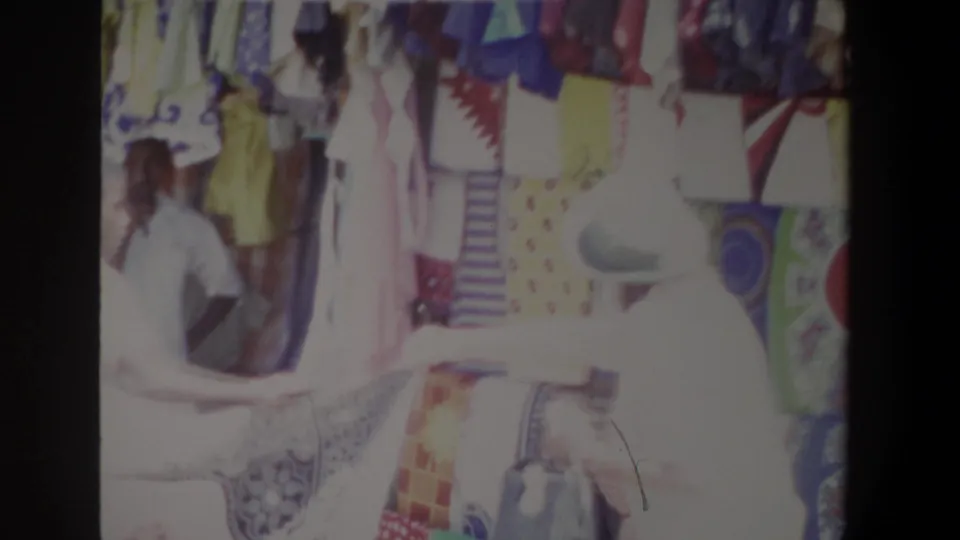In the Pretoria market clip, the light-hatted customer appears to handle or view patterned cloth while the seller stands behind the display.