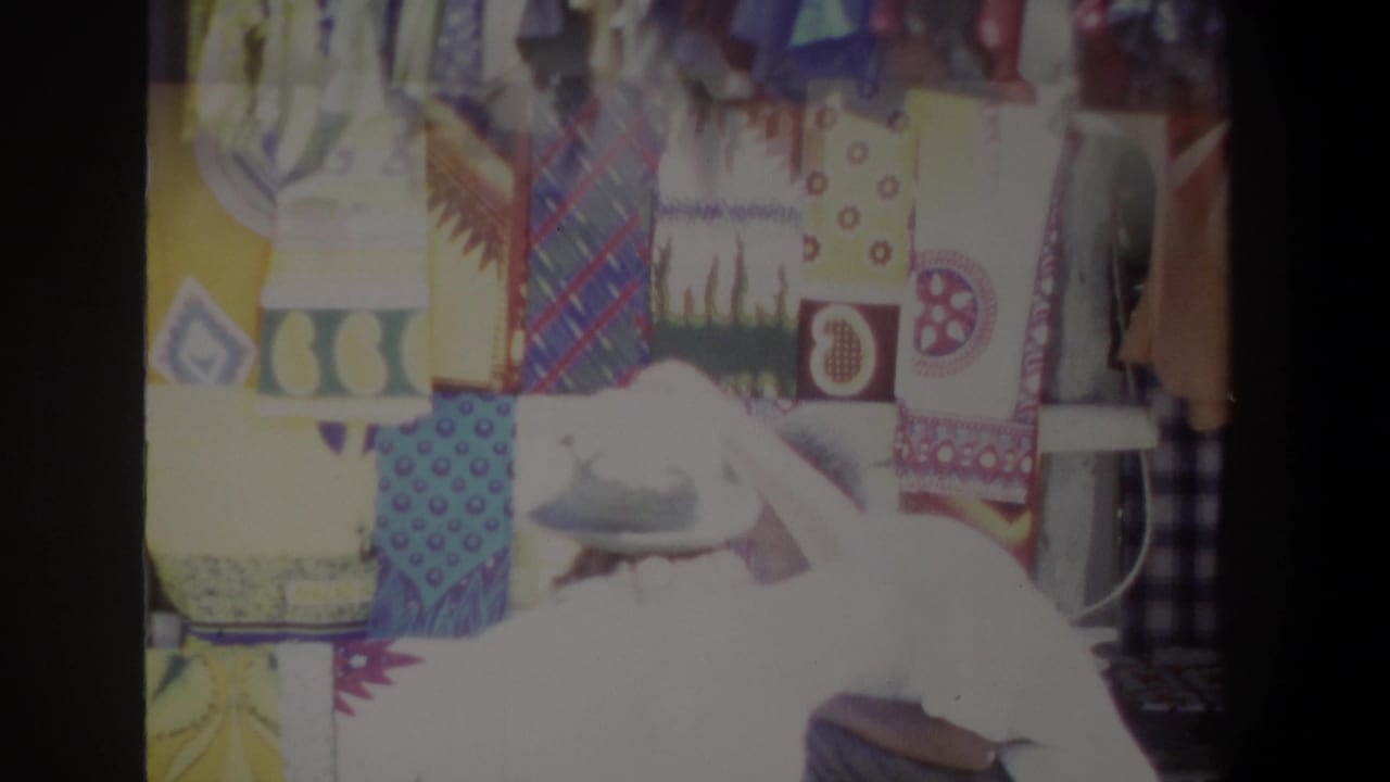 In 1969 Pretoria home-movie footage, a cap-wearing woman leans toward a bright textile display at an outdoor stall.