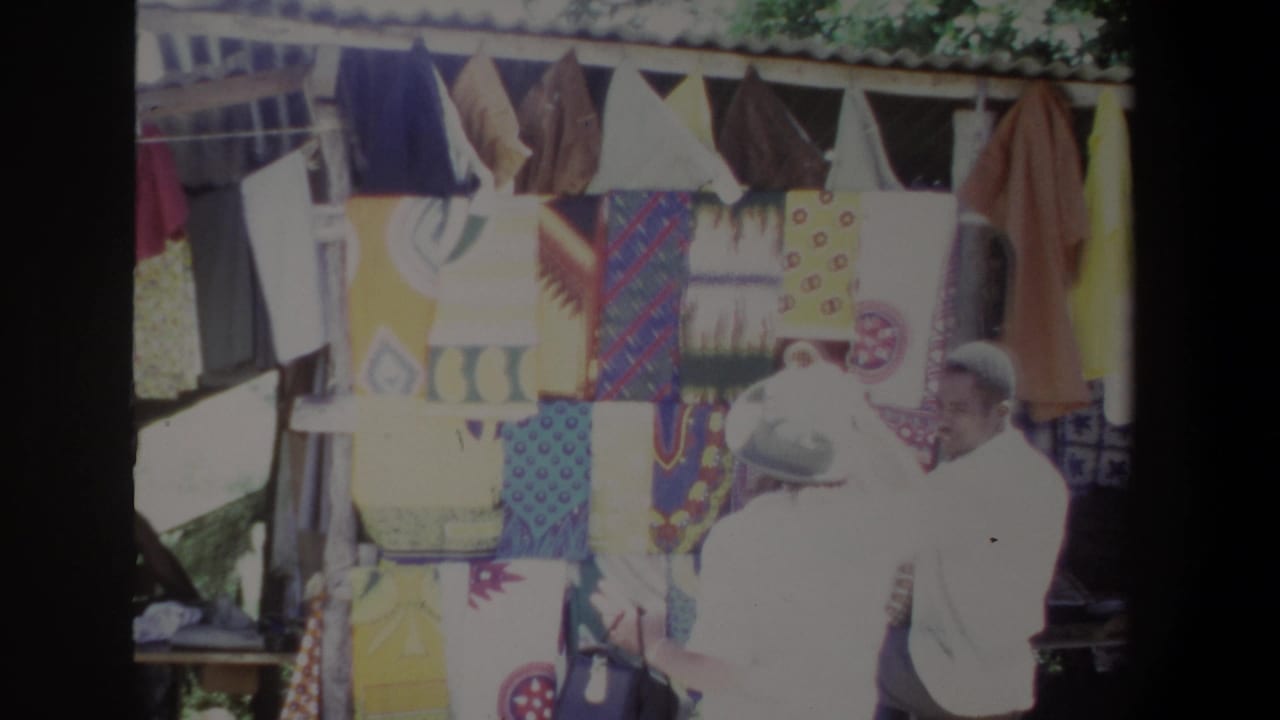 Vendor in a white shirt stands beside a textile display while a customer faces the stall.