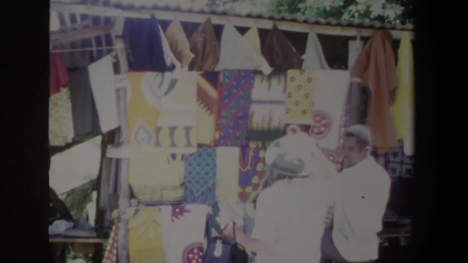 A man in a white shirt stands near a woman browsing colorful fabrics at an outdoor Pretoria clothing stall.