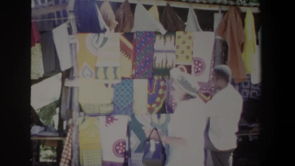 At a Pretoria outdoor stall, the vendor appears to show fabric to a cap-wearing visitor amid bold patterned textiles.
