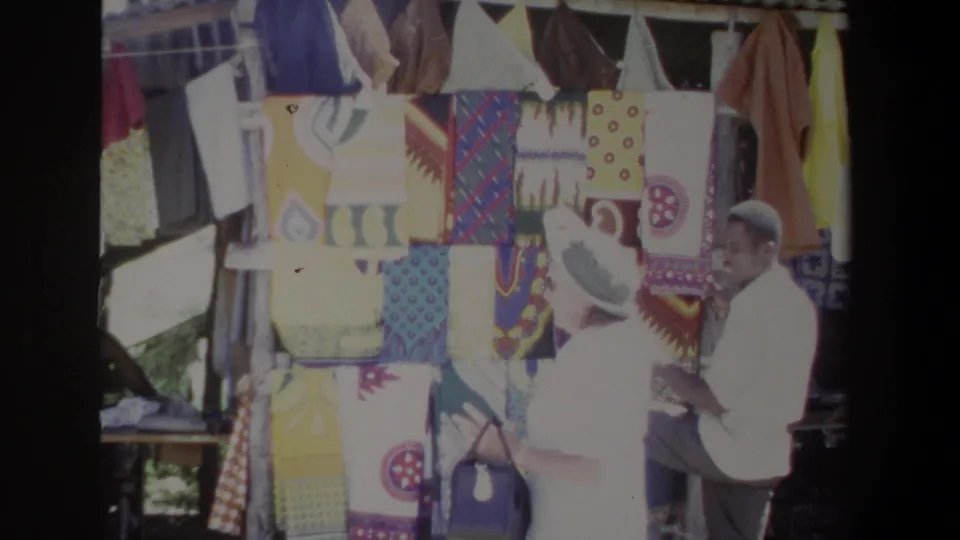 The 1969 Pretoria market scene shows a visitor and vendor interacting beside a dense display of patterned textiles.
