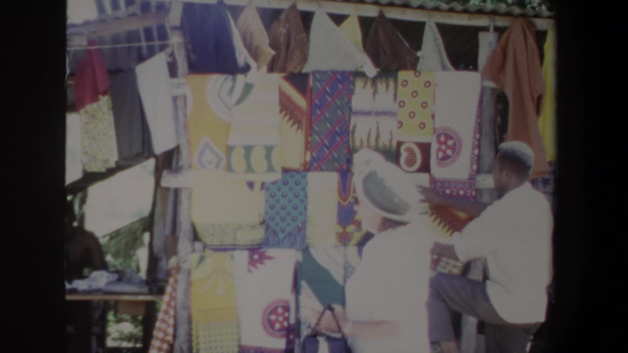 1969 Pretoria market frame showing colorful patterned blankets displayed at a stall, with a man seated beside the textiles.