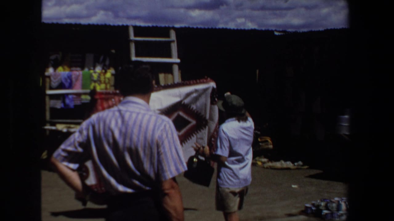 Two adults look at a patterned blanket outside a shaded market stall.