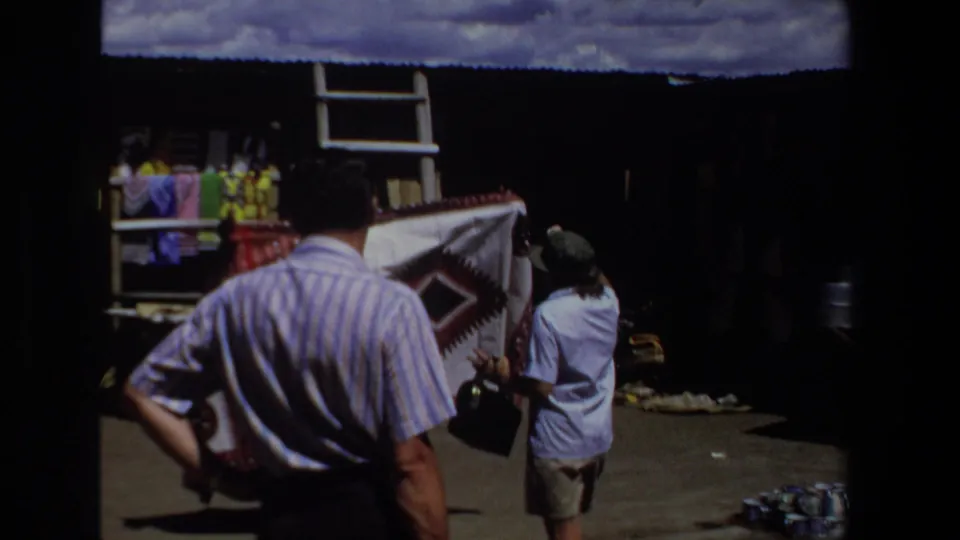 1969 Pretoria market view with an adult shopper and vendor examining a large patterned blanket near hanging textiles.