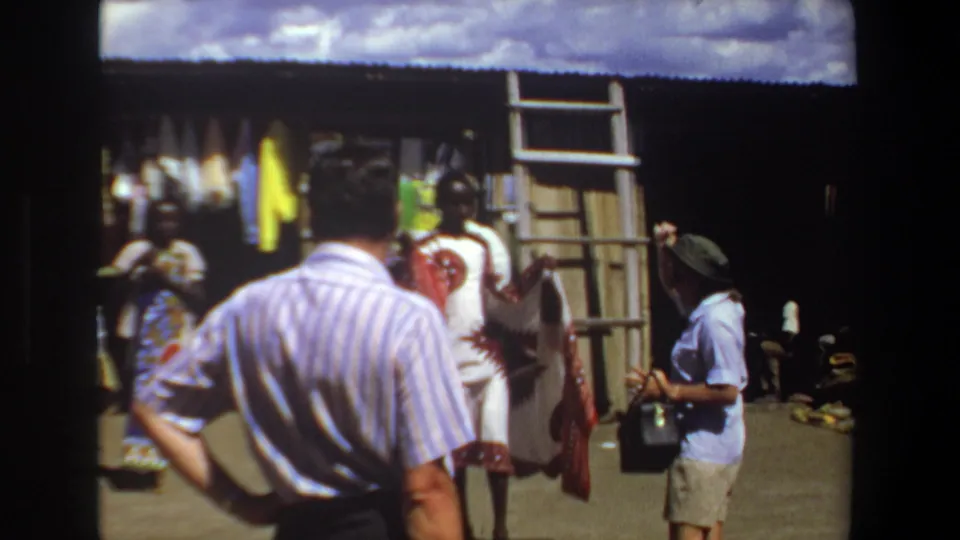 1969 Pretoria market frame showing a shopper from behind and a vendor near patterned blankets, hanging goods, and a wooden ladder.