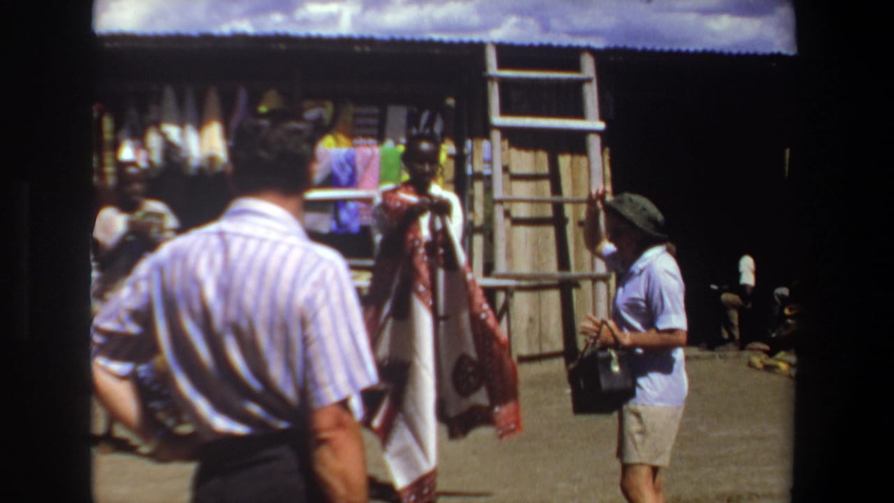 1969 Pretoria home-movie frame showing tourists near a vendor holding patterned textiles beside a shaded market stall.
