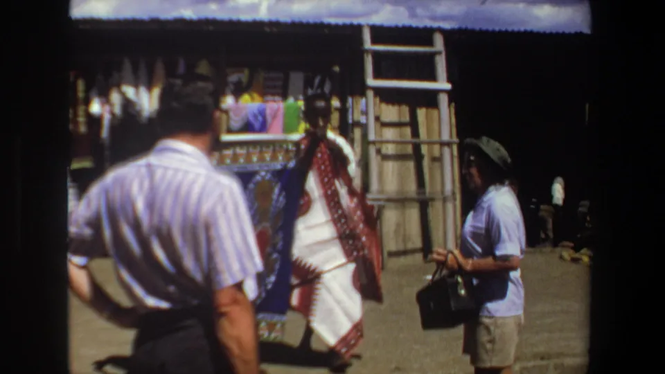 1969 Pretoria home-movie frame showing a textile vendor with fabric as tourists observe in a market setting.