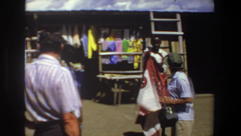 1969 Pretoria market frame with a vendor in patterned garments and a tourist woman near colorful hanging fabrics.