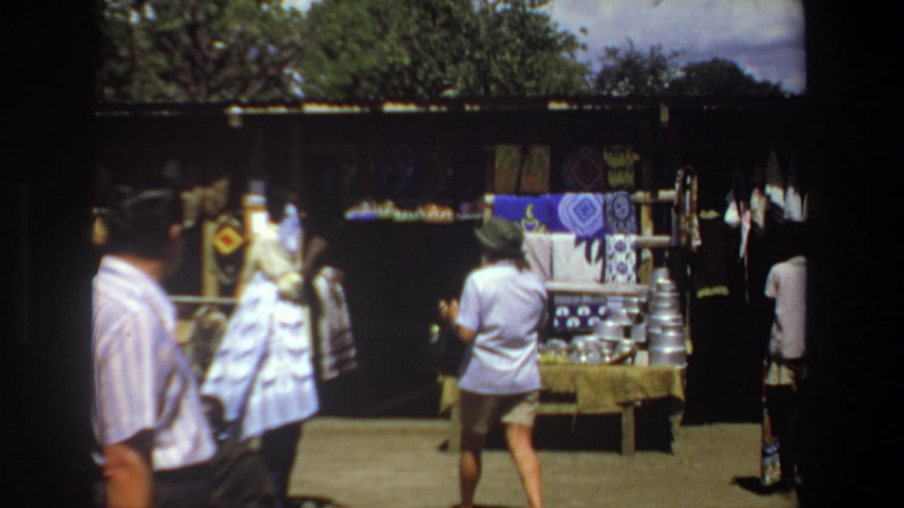 Shoppers stand before a market stall displaying fabrics and stacked metal wares.