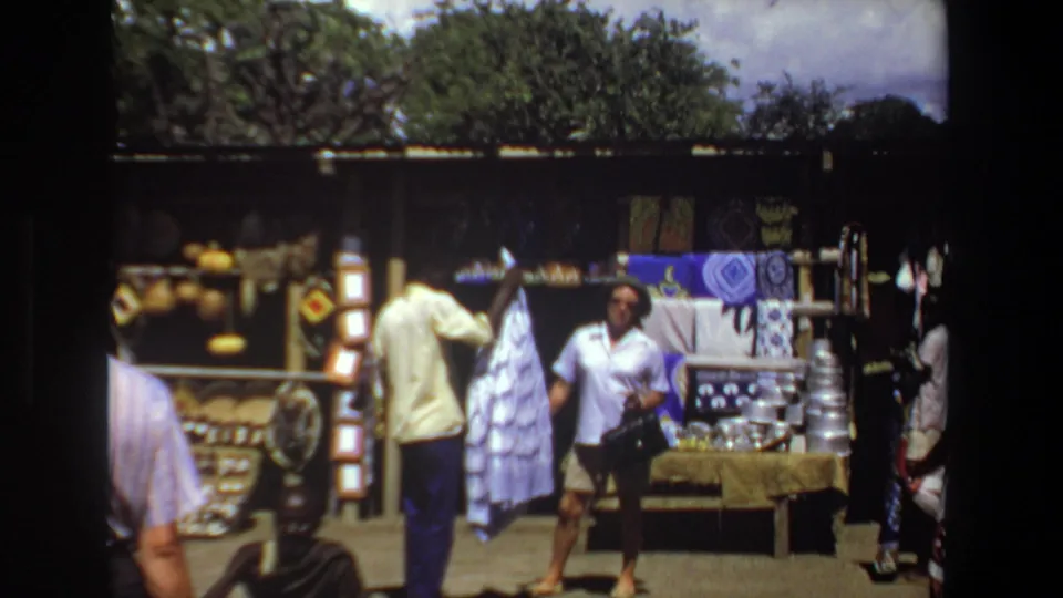 1969 Pretoria market frame showing a man handling a hanging cloth near a woman and a stall of textiles and metal wares.