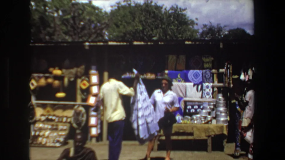 Pretoria, South Africa home-movie frame from 1969 showing adult shoppers near woven baskets and patterned textiles at an outdoor market stall.