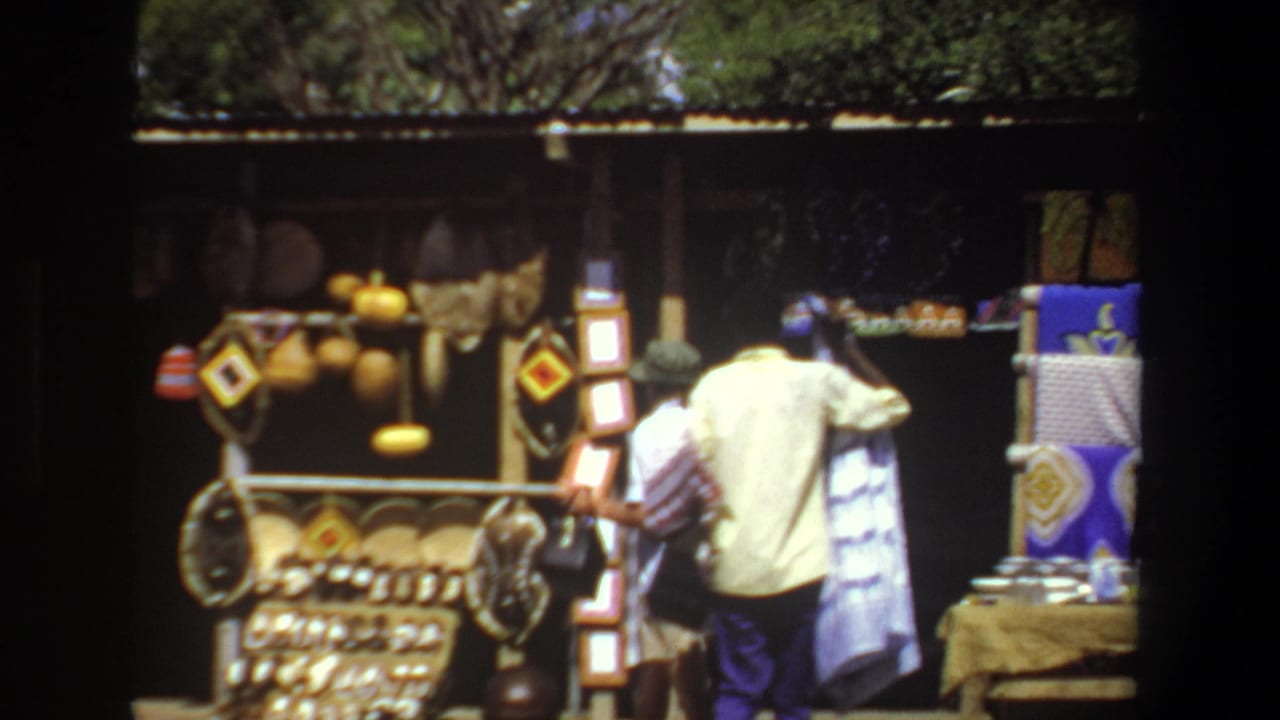 A man in a light shirt browses hanging baskets and patterned cloths at a Pretoria outdoor market stall in 1969 home-movie footage.