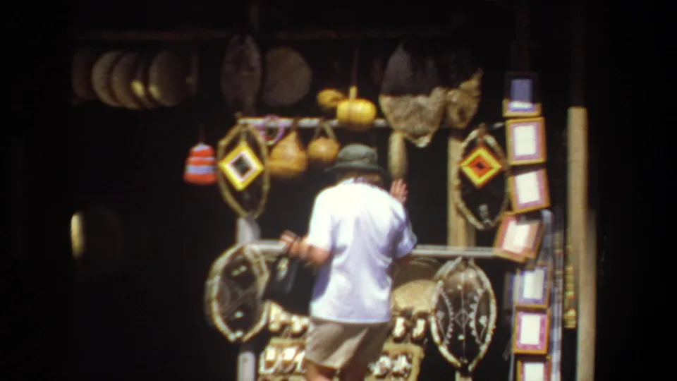 1969 Pretoria market frame showing an adult shopper from behind facing a stall with baskets, mats, and hanging handmade goods.