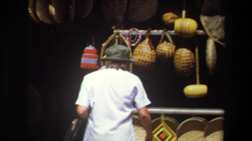 A man wearing a hat walks past hanging woven baskets and market goods in 1969 Pretoria travel home-movie footage.