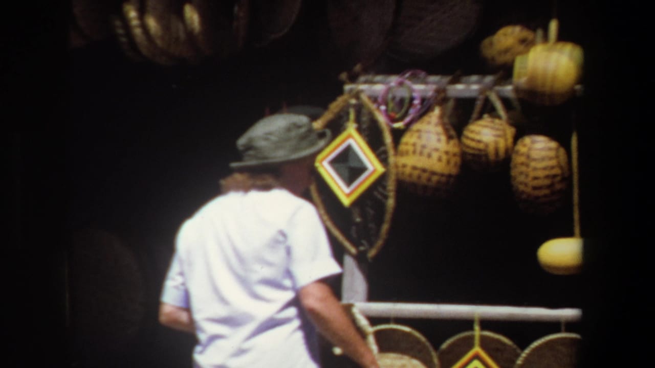 In Pretoria, South Africa in 1969, a woman in a white shirt and broad hat browses woven baskets, gourds and framed crafts at a market stall.