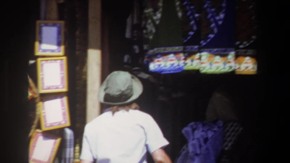 In the Pretoria market footage, the woman is seen from behind as she approaches a dim craft display with colorful textiles and framed objects.