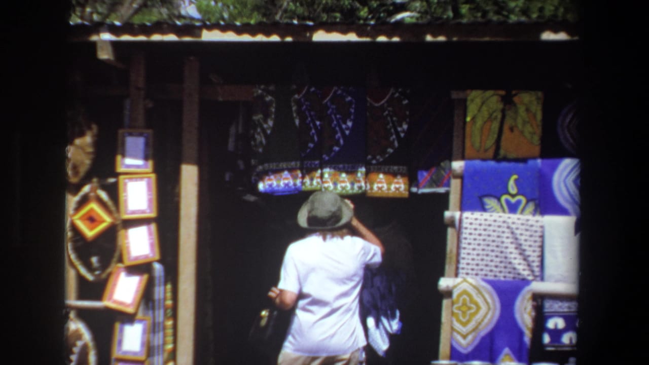 In Pretoria, South Africa in 1969, an adult woman in a white shirt and hat stands before a shaded stall with textiles and framed items.