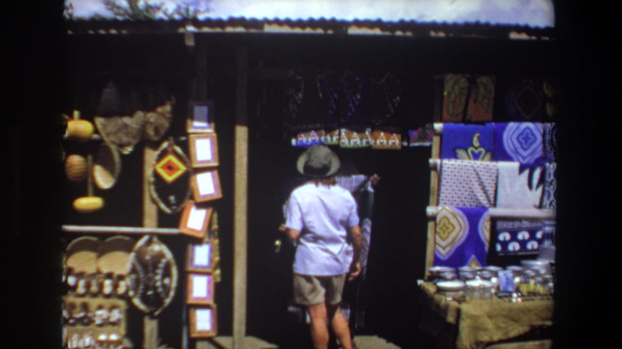 The 1969 Pretoria home-movie frame shows a woman seen from behind examining goods at a shaded gift stall with baskets and bright textiles.