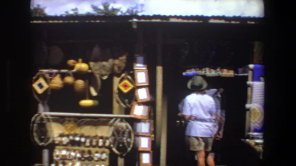 A woman in a hat and shorts moves along a Pretoria souvenir stall with woven items, framed pieces, and colorful textiles visible nearby.