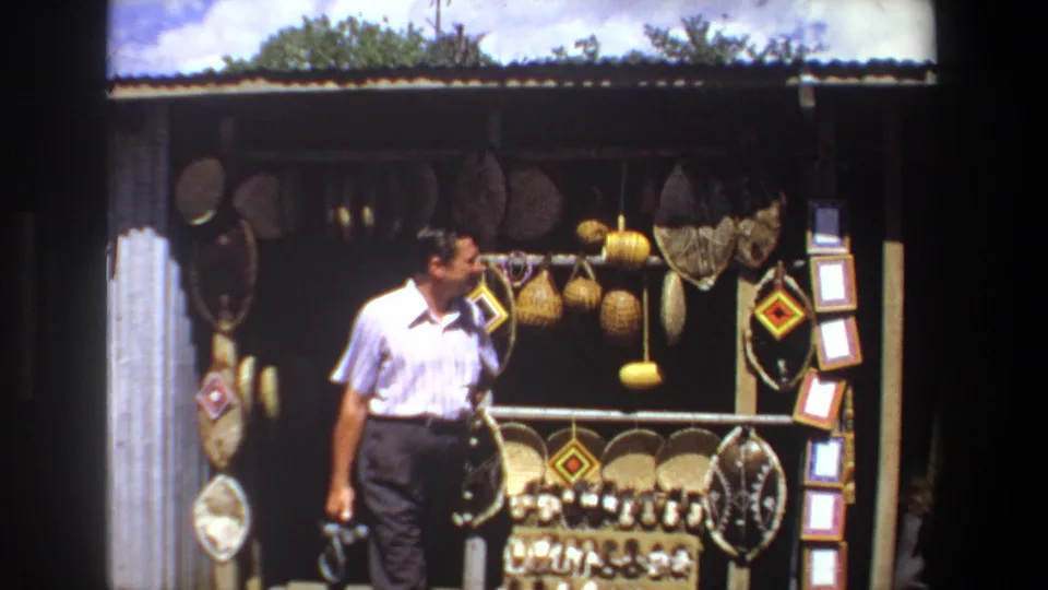 In the final 1969 Pretoria frame, an adult man in a white shirt stands in front of a shaded souvenir stall with hanging baskets and craft displays.