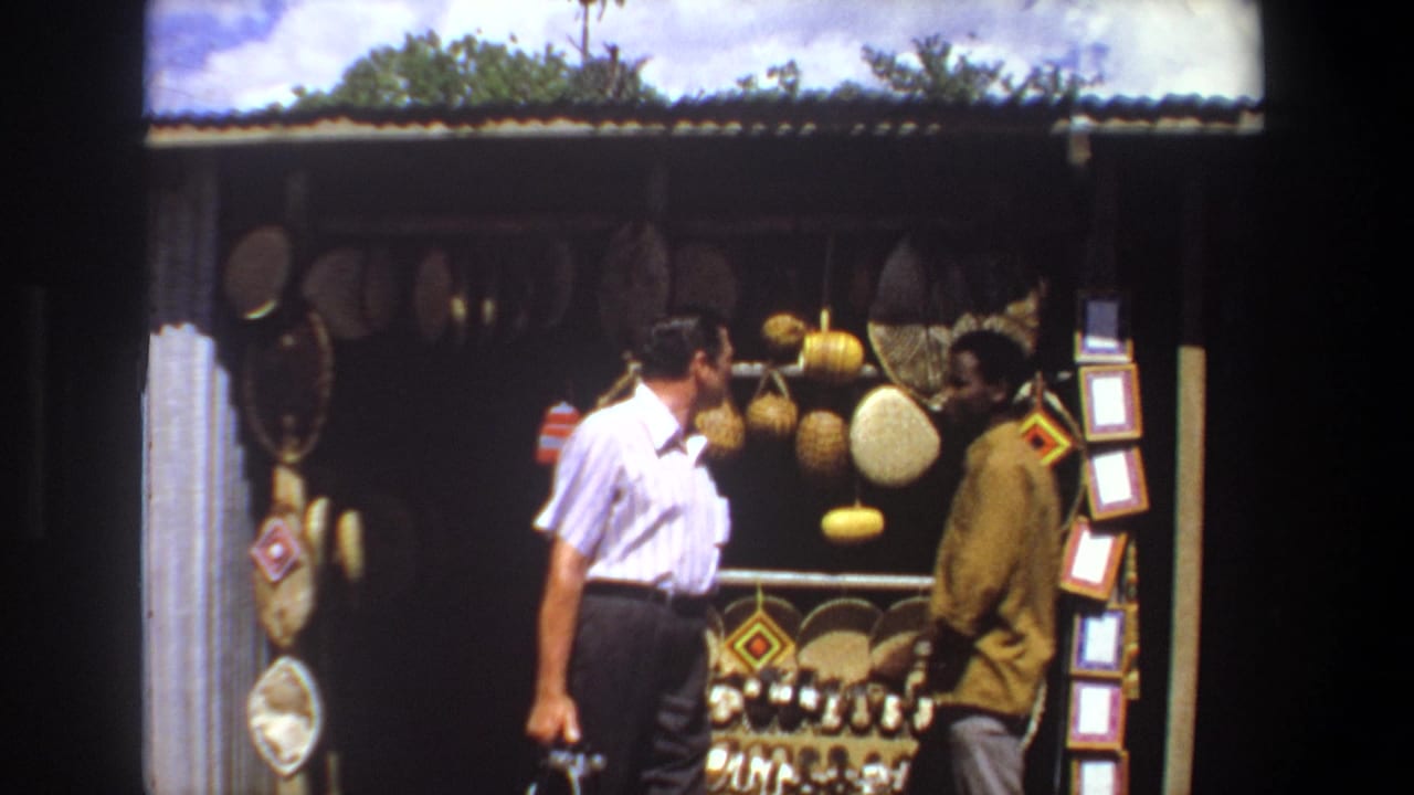 In 1969 Pretoria footage, a striped-shirt man looks toward a market stall displaying baskets, shields, and small framed items.