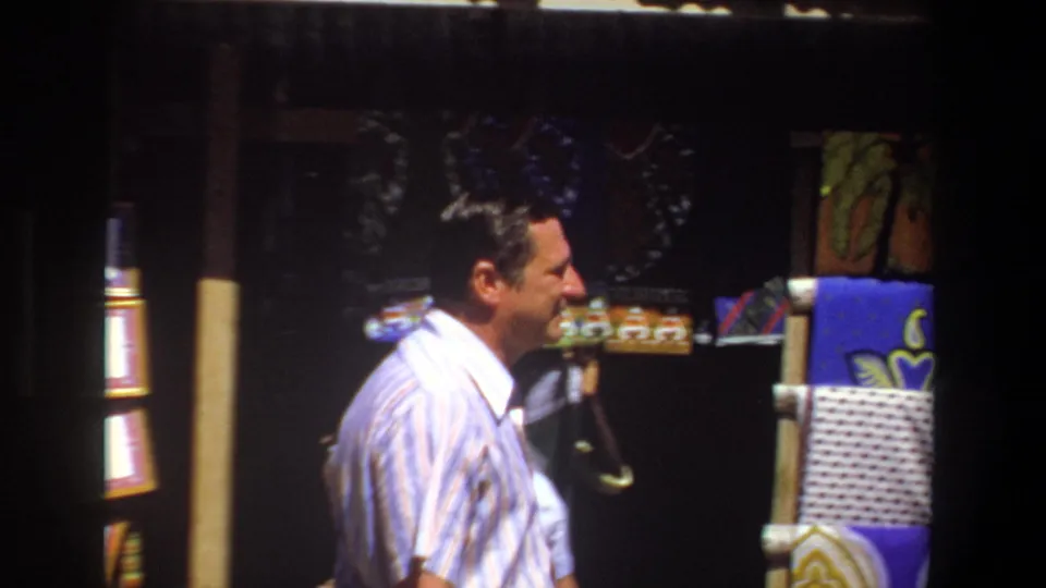 A man in a striped shirt smiles while walking past patterned textiles and craft goods in the 1969 Pretoria market footage.