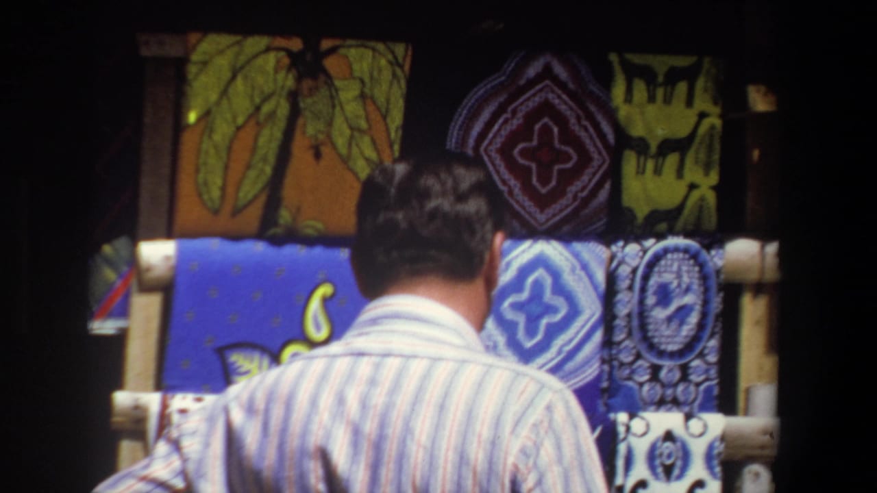 In 1969 Pretoria footage, a man in a striped short-sleeve shirt stands before a shaded display of colorful patterned textiles.