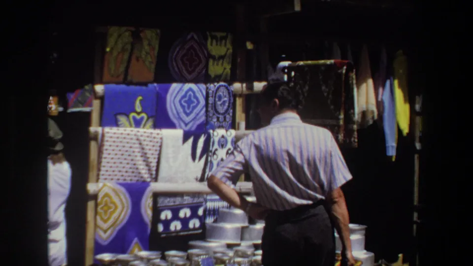 A rear view of the shopper shows him examining a fabric stall with patterned cloths above rows of shiny metal containers.
