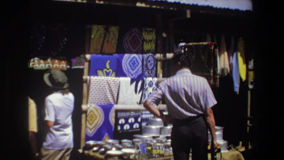 A woman in a hat passes or stands near the shaded Pretoria textile stall as the striped-shirt man looks at the display.