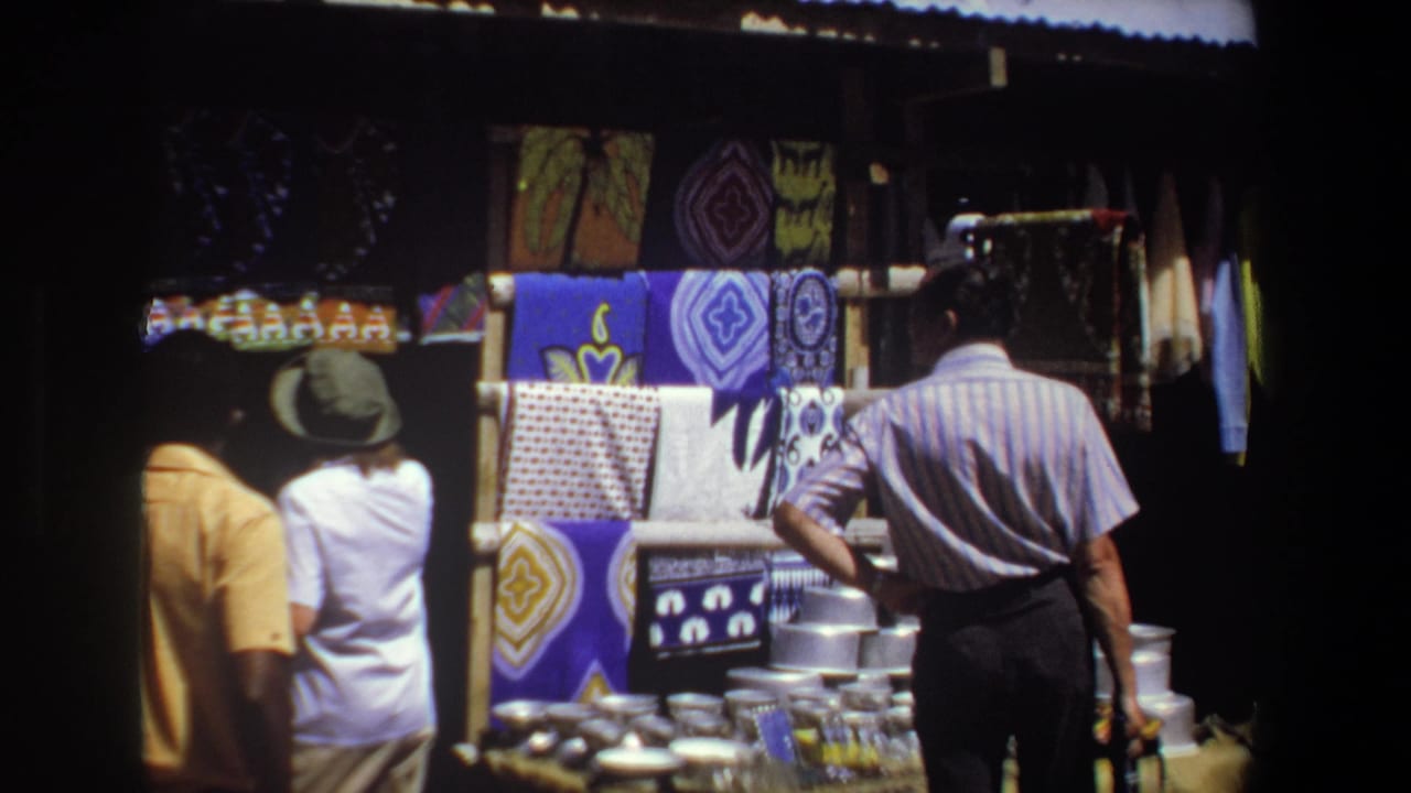 Pretoria, South Africa, 1969: shoppers seen from behind browse a shaded stall with patterned textiles and stacked metal goods.