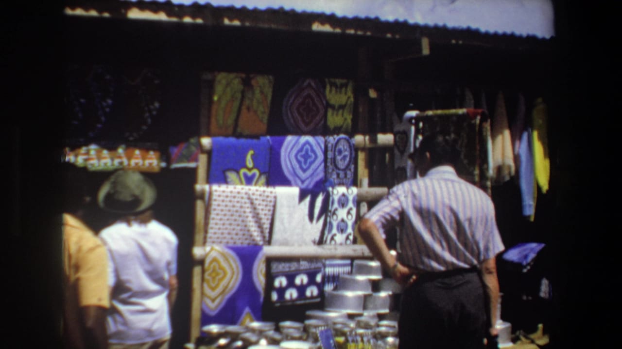 A man in a striped shirt faces a stall with bright fabrics and stacked metal wares.