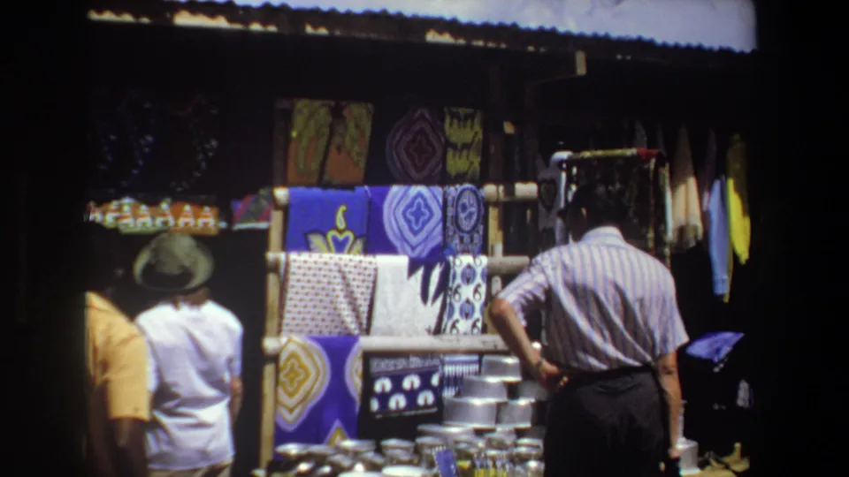 Pretoria, South Africa, 1969: a striped-shirted man and nearby shoppers look toward a display of textiles and metal goods.