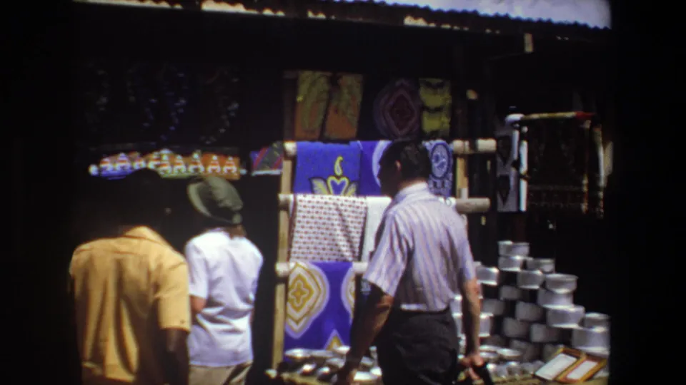 Pretoria, South Africa, 1969: shoppers pass a shaded stall where patterned fabric hangs above stacks of metal containers.