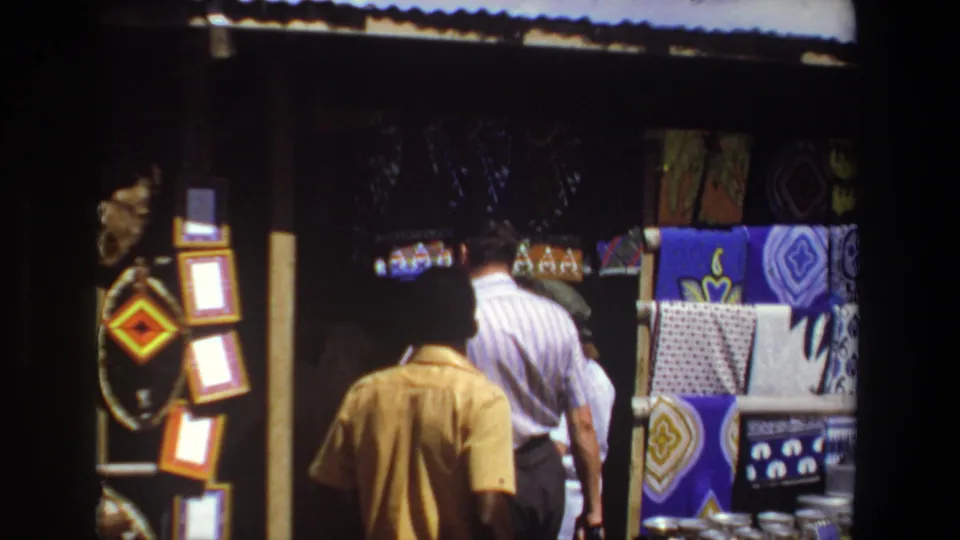 Pretoria, South Africa, 1969: shoppers move along a market stall with vivid textiles and small framed items visible at the side.