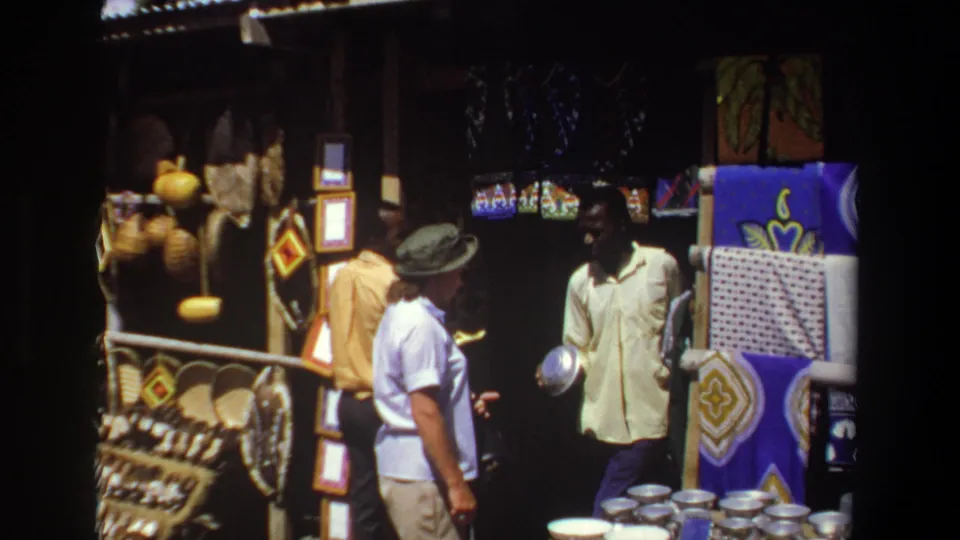 In the 1969 Pretoria home-movie footage, the woman gestures while the vendor stands among craft displays and stacked metal bowls.