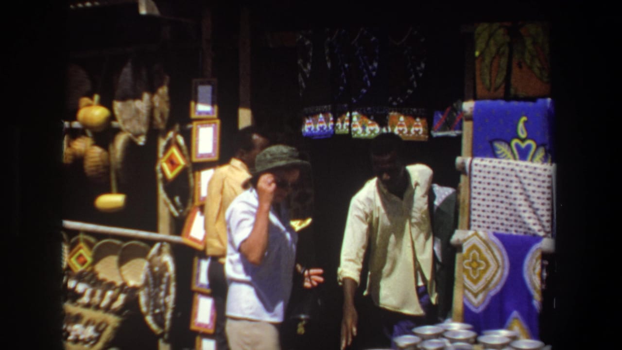 In 1969 Pretoria, a white man in a green bucket hat faces a Black vendor beside a stall displaying textiles, baskets, and framed crafts.