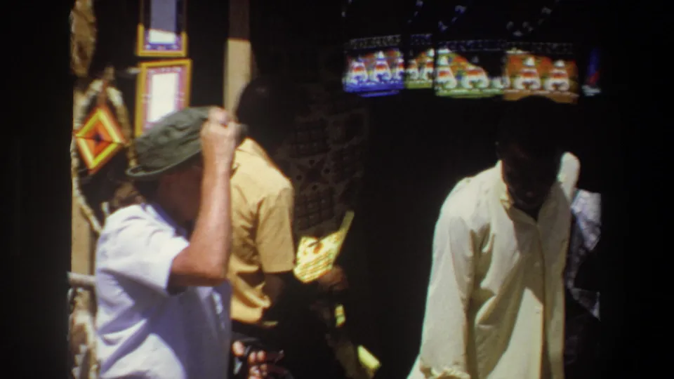 The handheld 8mm frame moves closer as the visitor in profile lifts his hand near his hat and the vendor stands partly shaded by the stall display.