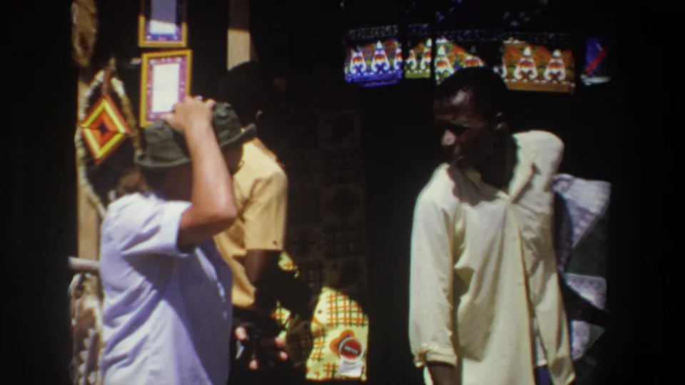 At the Pretoria craft stall, the white visitor stands with his hand near his hat while the Black vendor faces him beside patterned textiles and woven goods.