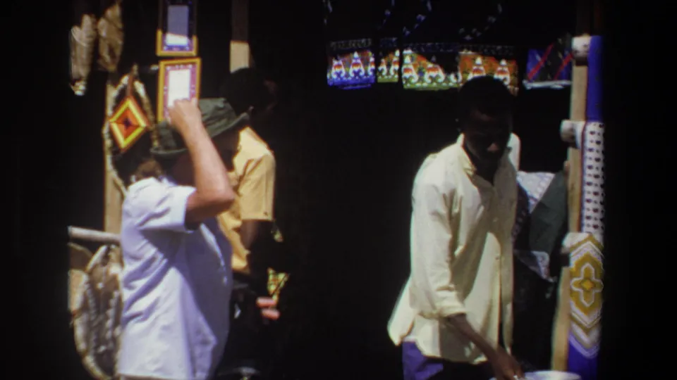 In 1969 Pretoria, a tourist woman in a green hat browses a shaded craft stall with a vendor nearby and patterned goods around them.