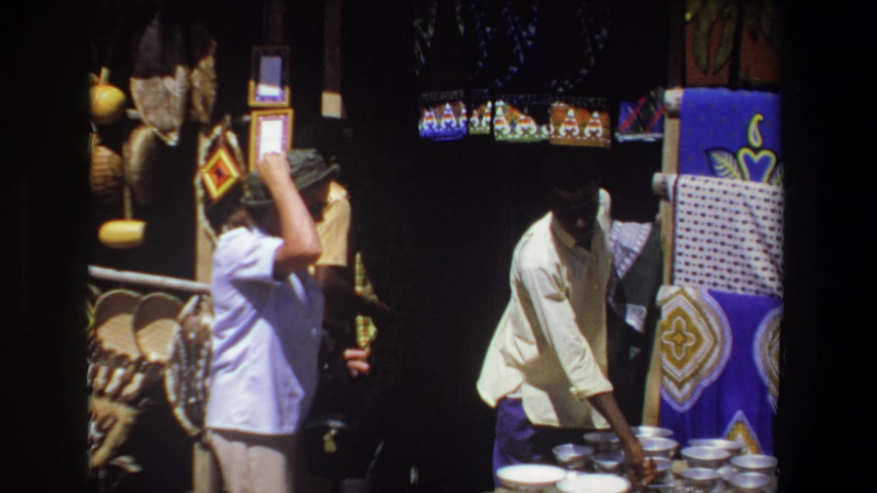 Vendor reaches toward bowls or small vessels beside colorful blankets at a market stall.