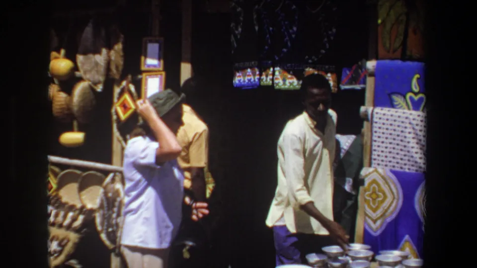 A 1969 Pretoria home-movie frame shows the tourist and vendor near baskets, patterned textiles, and small wares at a shaded stall.