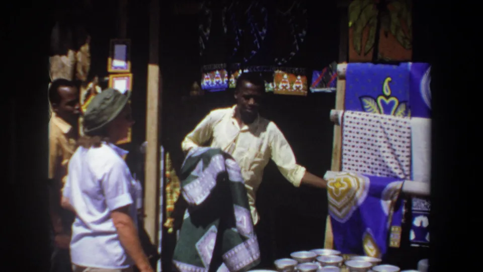 The vendor spreads a green patterned textile in front of the woman, with baskets, bowls, and colorful fabrics visible around the stall.