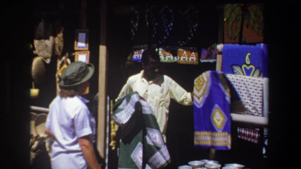 At the Pretoria stall, the vendor holds out folded cloth beside a blue patterned textile display as the woman continues watching.