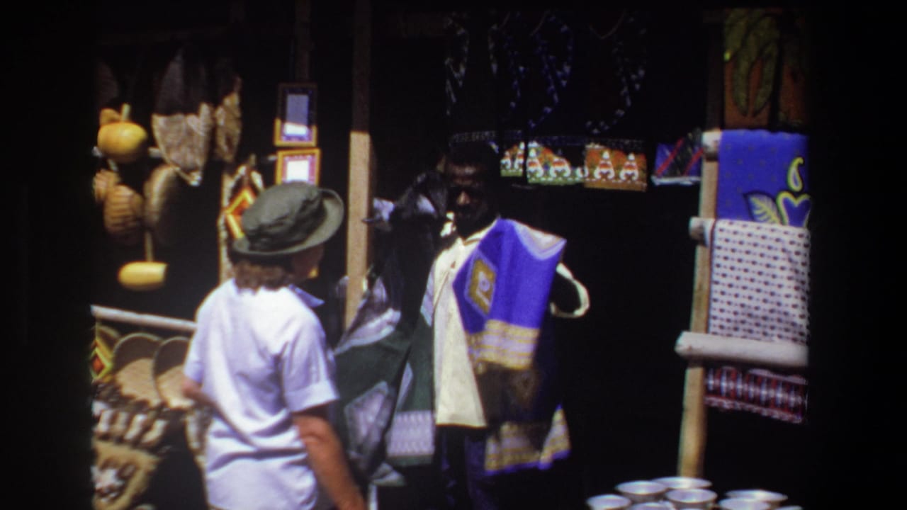In Pretoria, South Africa, 1969, a tourist in a hat faces a vendor displaying a purple patterned textile at a craft stall.