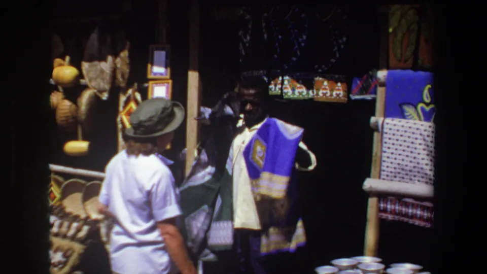 In Pretoria, South Africa, 1969, a tourist in a hat faces a vendor displaying a purple patterned textile at a craft stall.