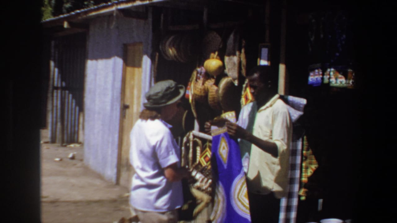 Vendor and tourist stand beside hanging baskets and textiles outside a small stall.