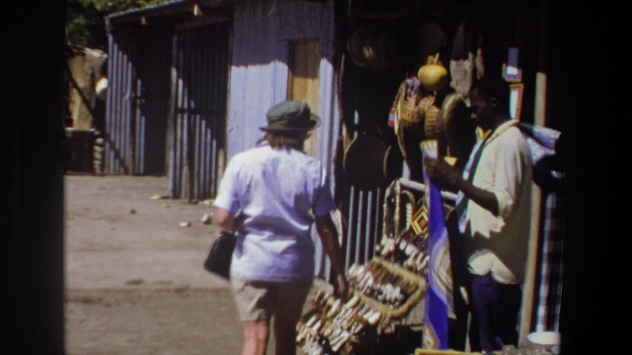 In supplied 1969 Pretoria footage, a tourist woman in a light shirt and hat walks toward a craft stall with baskets and hanging items.